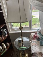 Full view of the floor lamp showing brass stand, glass shelf with a device on it, and white fabric lampshade near window.