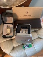 Photo of Seiko watch in the box, Jonathan Flagger leather wallet box, and Fossil travel clock metal tin case on beige chair