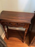 Front view of the solid wood night table showing the carved drawer front and turned legs on wooden floor against white wall.
