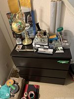 Photo of the brass desk lamp, black three-drawer cabinet with office supplies on top including notebooks, tape dispenser and rolls of plastic wrap, cordless phone, and miscellaneous small office items.