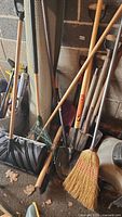 Photo showing assorted gardening tools leaned against a wall including snow shovel with black plastic scoop, wooden handled broom, and rakes