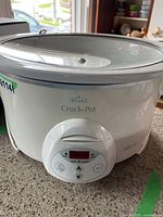 Front view of Rival Crock-Pot slow cooker showing white body, digital control panel, and glass lid on counter.