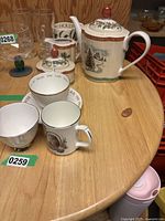 Photo showing the assortment of fine china teacups, mug with royal portraits, and Villeroy & Boch Festive Memories teapot with snowy winter scene on wooden table.