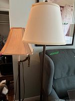 Two brass floor lamps with different shaped beige fabric shades near a blue wingback chair.