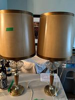 Two table lamps with gold bases, clear glass accents, and beige lampshades shown on a table, cords visible.