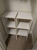 Two small white shelves with three compartments each, standing side by side in a corner.
