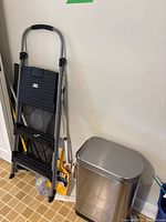 Step ladder leaning against wall with cleaning supplies and stainless steel garbage bin next to it.