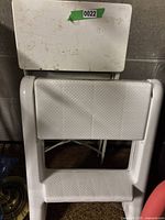 White plastic and metal folding step stool shown on side with textured steps and white top, visible dirt and wear
