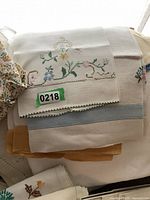 Photo showing a stack of folded tablecloths and napkins with floral embroidery and lace edges in whites and beiges.