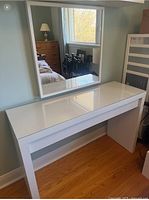 White modern vanity with attached large rectangular mirror, shown against bedroom wall, with hardwood floor.