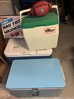 Three coolers stacked in basement: a vintage blue metal Coleman cooler, a white plastic Oscar by Coleman cooler with green lid, and a large white and blue plastic Coleman cooler. Football resting on top and boxed bag toss game behind.