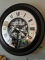 Front view of black-framed wall clock with Roman numerals and visible gears on mirrored face showing some room reflection.