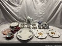 Full lot showing assorted porcelain and glassware set on white fabric background. Includes tea set, plates, jars, bowl, and sauce pot arranged in rows.