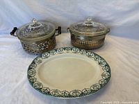 Two Pyrex covered servers with metal carriers and decorative glass lids positioned behind a Stratford patterned oval serving platter on a neutral background.