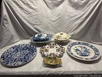 Wide shot showing all six ceramic serving pieces on a white cloth backdrop, including platters, covered dishes, and handled dishes, varying in size and color patterns, predominantly blue and white with some brown, yellow and gold.