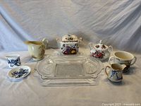 Seven-item lot on cloth backdrop including clear glass sandwich tray center, several ceramic creamers, sugar bowls, ashtray set, and Bunnykins mug.