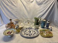 Overall lot showing assortment of figurines, pitchers, bowls, plates, and vases spread on white cloth background