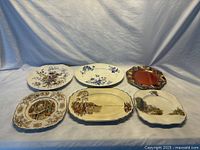 Overall group photo showing six assorted plates and serving platters, arranged on light blue fabric.