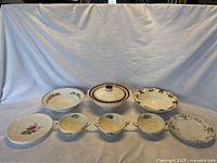 Full view of all 11 pieces of vintage dishware in cream and floral patterns including plates, bowls, and a covered serving dish, arranged on a white cloth background.