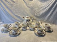 Overall view of teapot, creamer, sugar bowl, six cups and saucers arranged on draped backdrop.