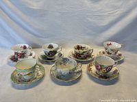 Front view showing seven fine bone china teacup and saucer sets with various floral and emblem patterns arranged in two rows.