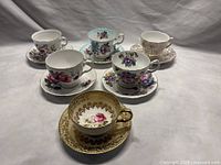 Six vintage teacup and saucer sets displayed together on neutral backdrop, showing various floral patterns and gold detailing.