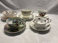 Five porcelain teacup and saucer sets displayed together showing various floral and pictorial designs with gold trim.
