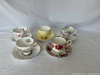 Image showing five porcelain teacup and saucer sets arranged in a semicircle on white background, each with distinct floral or gold rim designs.