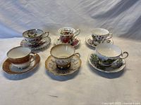 Six assorted vintage teacup and saucer sets arranged on white surface, showing various floral and gilded designs.