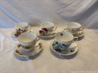 Five teacups and saucers with different floral patterns and gold trim, displayed on a white cloth background.
