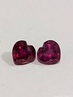 Two deep red heart-shaped rubies with facet cuts, positioned side by side on a white surface.