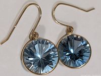 Pair of 10KT gold dangle earrings with round light blue faceted blue topaz stones, shown laid flat and angled to highlight facets and setting.