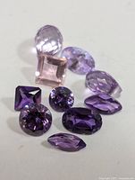 Photo showing multiple faceted loose genuine amethyst gemstones in various cuts and shades of purple, arranged on a white surface.