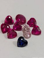 Image showing six deep pink heart-shaped CZ stones, one dark blue heart-shaped CZ stone, and two clear oval CZ stones arranged together on a white background.