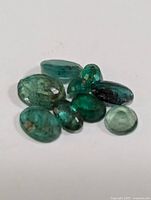Close-up image showing eight loose genuine emerald stones in various oval and round faceted cuts with natural inclusions and color variations.