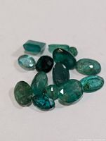 Close up photo showing 13 mixed cut loose genuine emerald gemstones in various green shades and shapes.