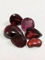 Photo showing six genuine garnet gemstones of mixed cuts including round faceted, pear-shaped faceted, and a polished cabochon with rich red hues.