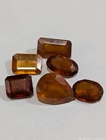 Five faceted hessonite garnet gemstones in amber to burnt orange hues and various cuts arranged on a white surface.