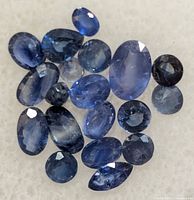 Close-up image of 18 faceted sapphire gemstones on a white background, various sizes and shapes showing deep blue color.