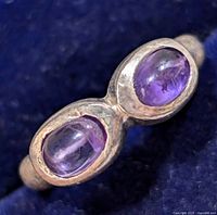Close-up image of silver ring showing two oval cabochon amethyst stones with natural inclusions, set side by side on a silver band