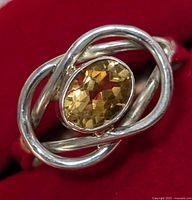 Close-up of silver ring with oval faceted citrine gemstone on intertwined silver band, displayed on red background.