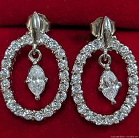 Front view of silver oval hoop earrings encrusted with round cubic zirconia stones and dangling marquise-cut stone inside hoops.
