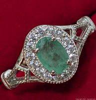 Close-up front view of silver ring featuring oval emerald and clear stone halo on red background.