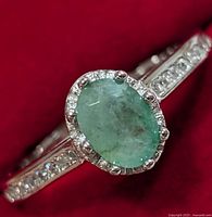 Close-up of the ring from the front showing the oval emerald surrounded by small clear stones and sparkling accents along the band