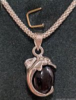 Silver necklace with woven chain and garnet dolphin pendant, laying on black fabric with a small bent metal item beside it.