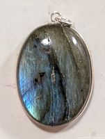 Close-up front view of an oval labradorite stone pendant with silvery blue iridescence and natural dark inclusions, mounted in a silver bezel with an attached loop.