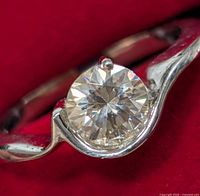 Close-up of silver ring with round moissanite gemstone on red background showing twisted band and prong setting.