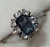 Close-up image of silver ring showing blue rectangular center stone with clear stones surrounding it in halo setting on silver band.
