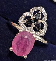 Close-up of silver ring featuring oval ruby center stone and sapphire accents on dark blue background