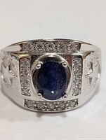Close-up image of the front of the silver ring showing oval blue sapphire set in the center, surrounded by small clear stones and polished silver band.